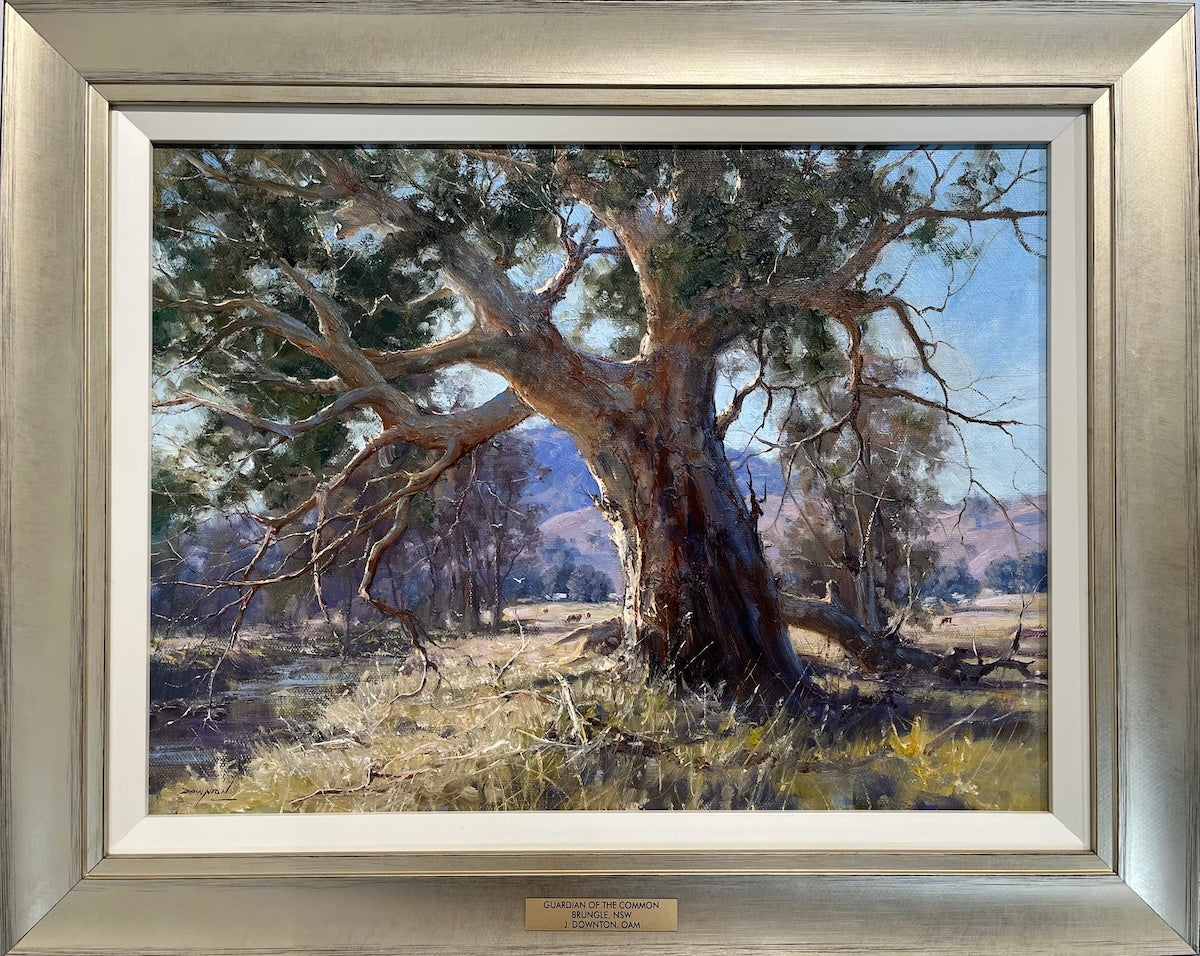 A large old gum tree on the banks of a river, with cows grazing, a homestead and hills in the distance. Framed view.