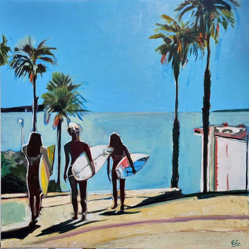 Three girls, with surfboards under their arms, heading towards the ocean for a surf, down a path lined with palm trees. There is a headland in the distance, across the water.