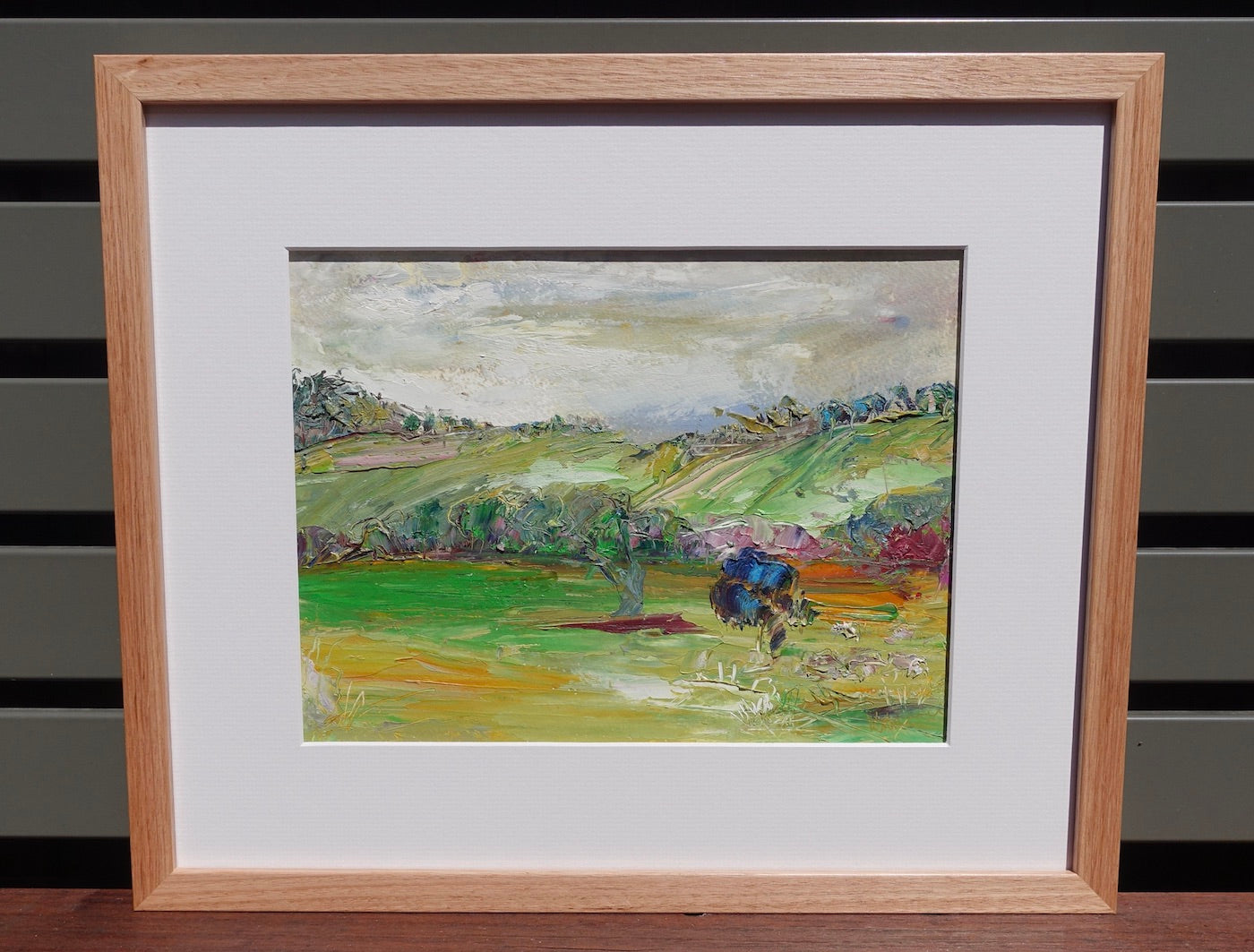 Abstract painting of a green countryside with a blue tree in the foreground. Framed view.