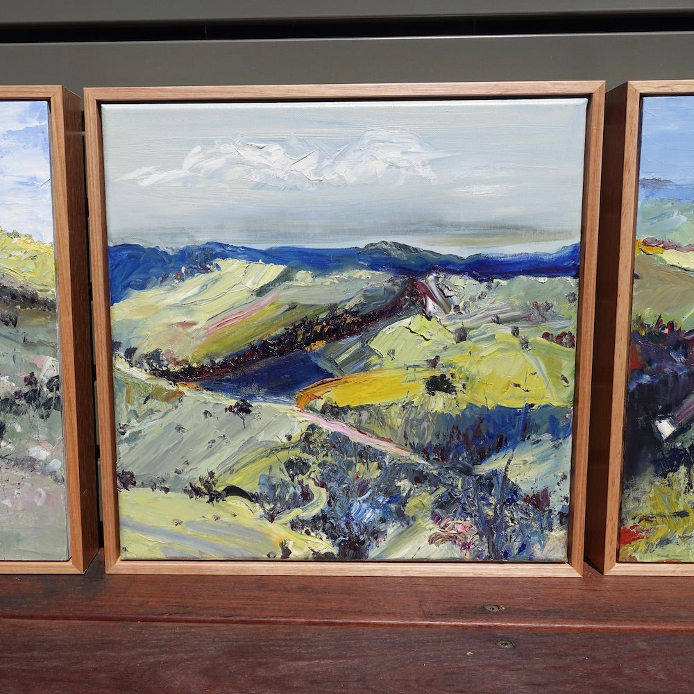 Abstract oil painting of a country landscape with steep hills and lots of colour, yellow, pink, green and blue against a pale blue sky with white clouds. Shown next to other paintings.