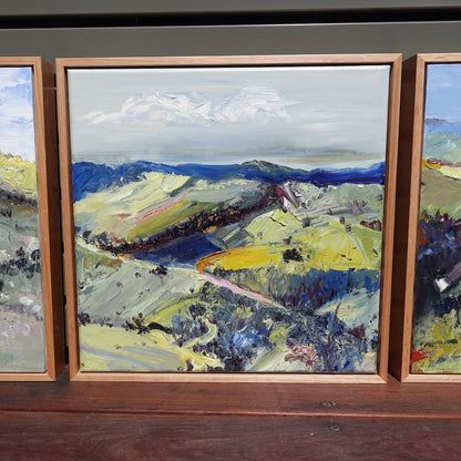 Abstract oil painting of a country landscape with steep hills and lots of colour, yellow, pink, green and blue against a pale blue sky with white clouds. Shown next to other paintings.