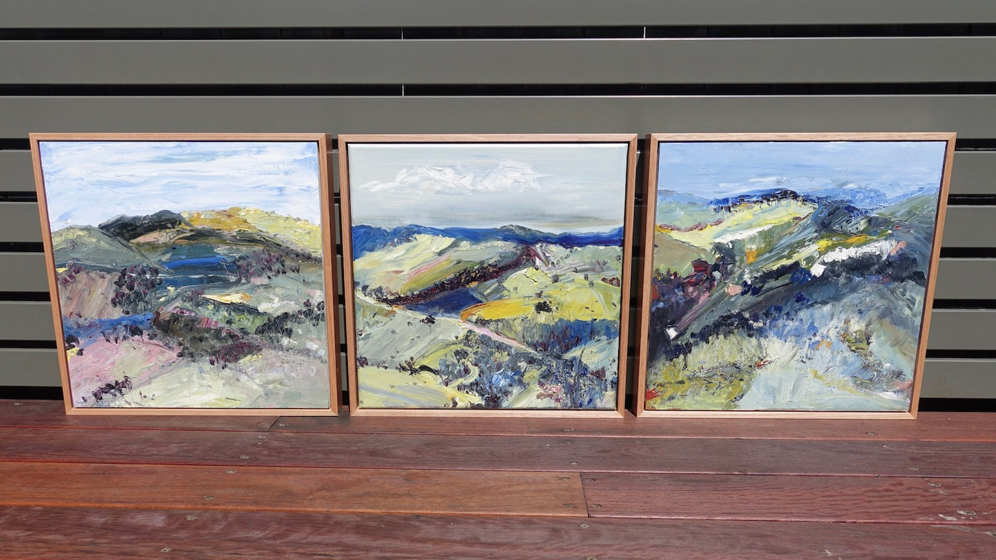 Abstract oil painting of a country landscape with steep hills and lots of colour, yellow, pink, green and blue against a pale blue sky with white clouds. Shown next to 2 matching paintings.