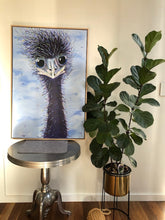 Load image into Gallery viewer, A painting of an emu with blue ringed eyes against a blue and white cloudy sky. Shown in situ against a wall, on a small round  metal table.

