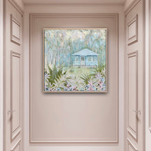 Load image into Gallery viewer, An idyllic pale blue beach cottage with white trim on the windows and verandah. The house is surrounded by palm trees, with a white puppy on the grass in front of the cottage and a garden with fern fronds and flowers. Shown in situ on a beige pink, hallway wall.

