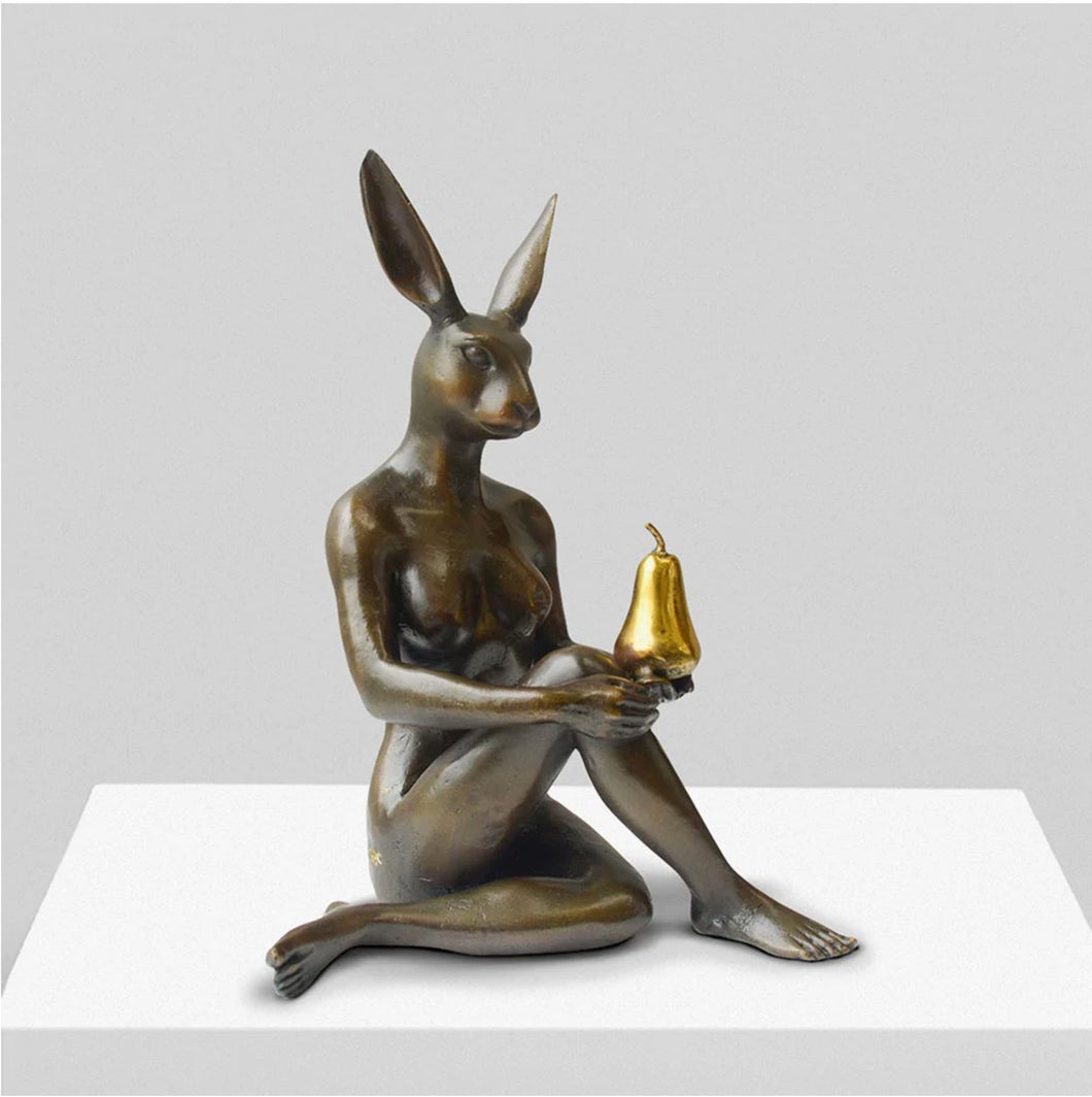 Bronze sculpture with gold patina of rabbit woman sitting on the floor with her legs crossed holding a golden pear.