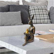 Load image into Gallery viewer, Bronze sculpture with gold patina of rabbit woman sitting on the floor with her legs crossed holding a golden pear. In situ sitting on a marble table.
