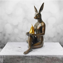 Load image into Gallery viewer, Bronze sculpture with gold patina of rabbit woman sitting on the floor with her legs crossed holding a golden pear. In situ, sitting on a piece of concrete.
