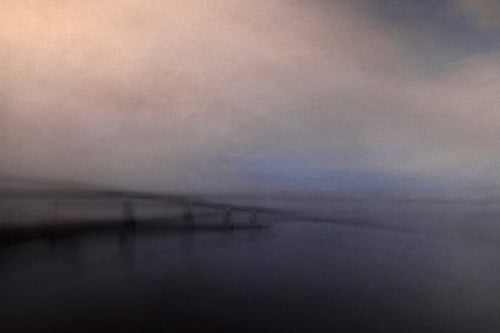 A misty view of a bridge over the ocean with a faint patch of blue showing in the sky.