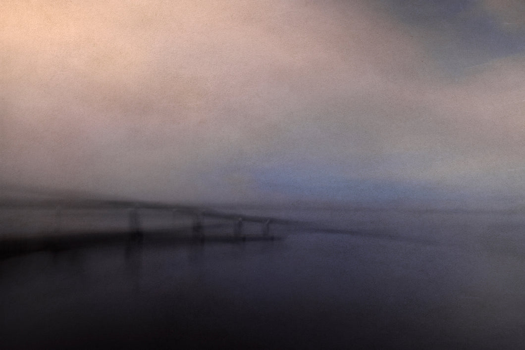 A misty view of a bridge over the ocean with a faint patch of blue showing in the sky.