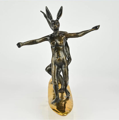 Bronze sculpture of a dog man and rabbit woman on a surfboard with gold patina. Front view.