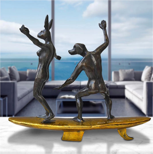 Bronze sculpture of a dog man and rabbit woman on a surfboard with gold patina. In situ on a marble table.