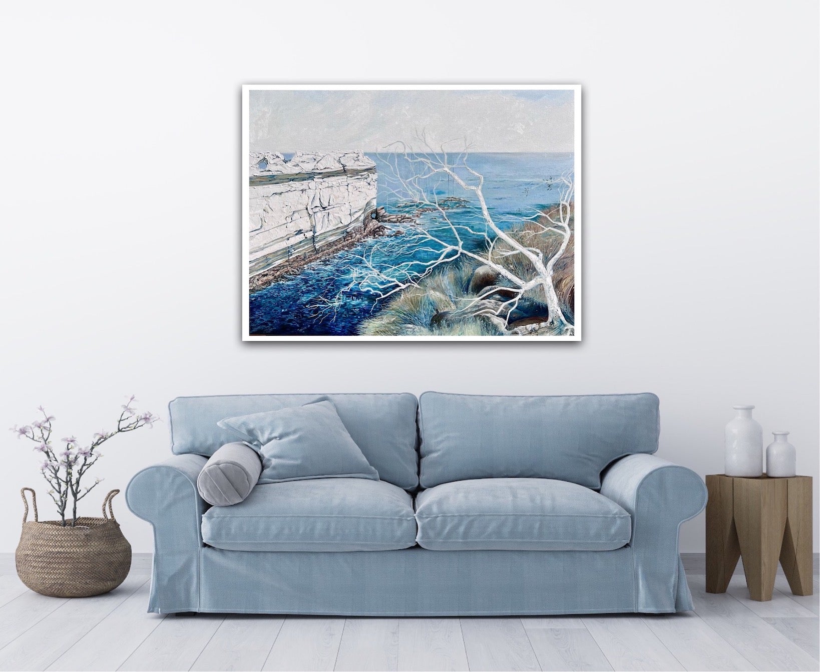 Abstract painting showing a headland jutting out into the ocean, in shades of blue and white. Shown on a white wall above a pale blue sofa.